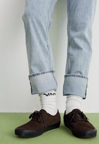Rudi zomšinių sportbačių su juodais padais, derinančių su sukneigtomis šviesiai mėlynomis džinsomis ir baltais kojinėmis, ant kurių matosi "VANS" logotipas.