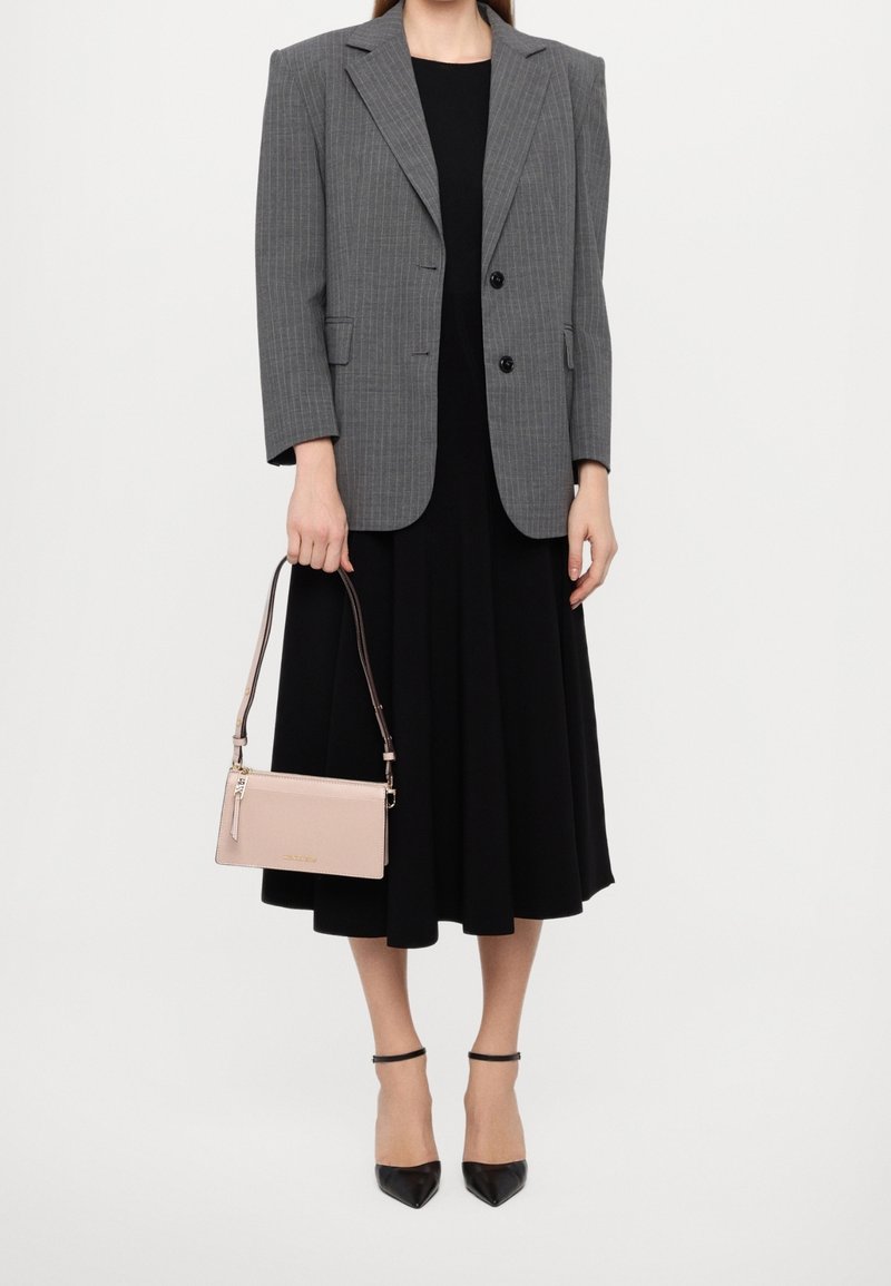 Personne portant un blazer gris à fines rayures sur une robe midi noire, tenant un sac à main rose pâle et portant des talons noirs à bride cheville.