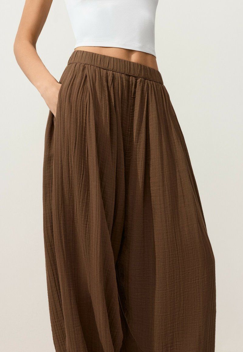 Femme portant un pantalon marron texturé ample, taille haute avec poches latérales et un haut court blanc sans manches.