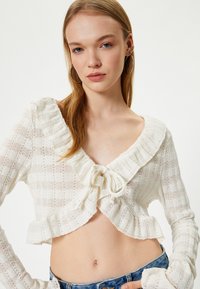 Weißer, verkürzter Cardigan mit einem Rüschenausschnitt, knopfartigem Verschluss und strukturiertem Strickmuster. Lange Ärmel mit fransigen Bündchen.