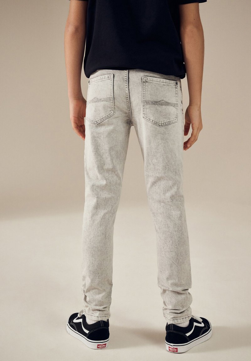 Hellgraue Jeans mit schlanker Passform, dezentem Used-Look und zwei Gesäßtaschen. Kombiniert mit schwarzen Sneakern mit weißen Akzenten.