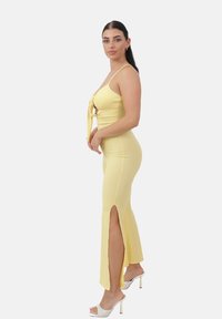 Vestido largo amarillo con una silueta ajustada, tirantes ajustables, detalle de lazo en el frente y aberturas laterales hasta el muslo. Acompañado de tacones transparentes.