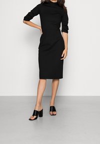 Robe noire ajustée à manches trois quarts, col haut et plis subtils ; associée à des sandales noires à talons carrés. Texture lisse.