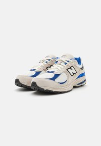 Chaussures de sport New Balance présentant un mélange de filet blanc et de daim beige, rehaussées de détails bleus et noirs, avec une semelle en caoutchouc texturé.