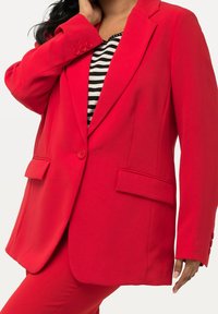 Femme portant un blazer rouge vif sur mesure et un pantalon assorti, avec un haut rayé noir et blanc, main près du cou.