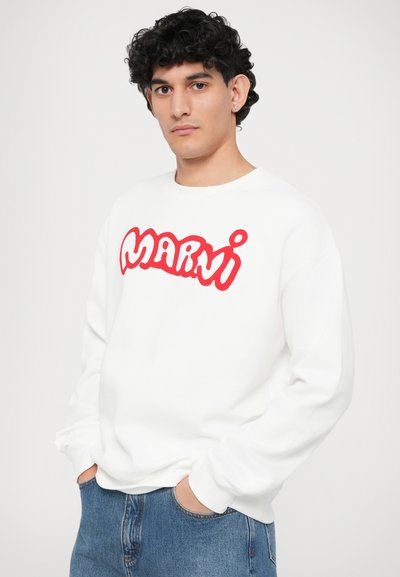 Sudadera blanca con logo estilizado "Marni" en rojo, cuello redondo y puños acanalados, combinada con vaqueros azules de corte relajado.