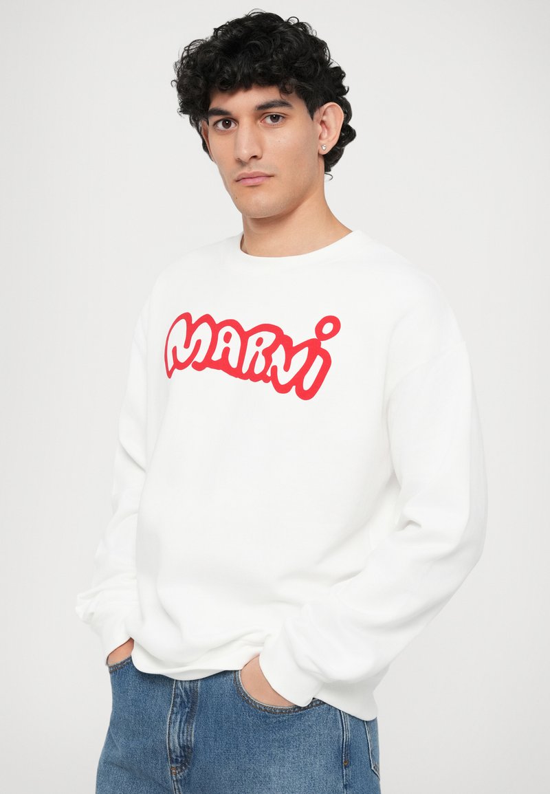 Vit sweatshirt med röd stiliserad "Marni"-logotyp, rund hals, och ribbade muddar, bärs med blå jeans som har en avslappnad passform.