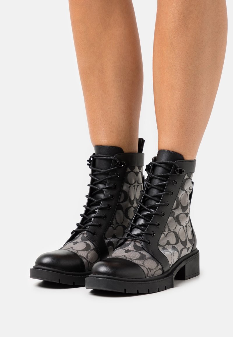 Bottines noires et grises à lacets avec côtés en tissu à motifs portées sur des jambes nues sur un fond clair.