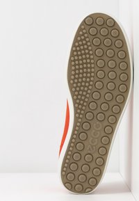 Suela de goma con patrones de agarre circulares en marrón, con una parte superior de ante naranja con detalles en blanco y de marca.