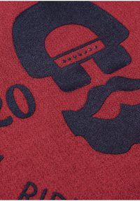 Roter Stoff mit einem großen, dunklen grafischen Design. Das Design beinhaltet eine stilisierte Helmform und ein wellenartiges Element mit einer strukturierten Oberfläche.