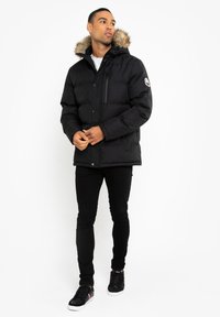Sort pufferjakke med en hætte i pels, lynlås og trykknaplukning samt en sidelomme. Modellen har sorte bukser og sneakers på.