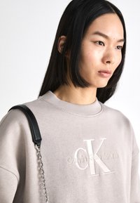 Svijetlosmeđa dukserica s rebrastim ovratnikom i izvezenim logotipom "Calvin Klein Jeans" u bijeloj boji. Crni remen s detaljima od lančića.