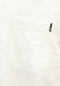 Λευκό υφασμάτινο υλικό με υφή και μια μικρή τετράγωνη τσέπη που φέρει μια μαύρη ετικέτα με λευκό λογότυπο δέντρου στην επάνω δεξιά πλευρά.