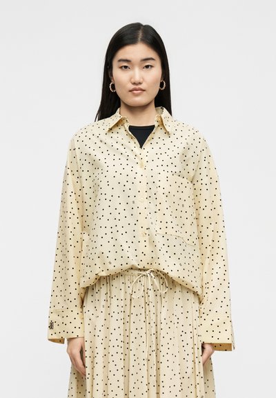 Femme aux longs cheveux noirs portant un chemisier beige et une jupe à pois noirs, debout devant un fond clair uni.