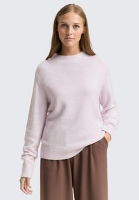 Pulover lung cu mânecă lungă, de culoare roz pal, cu decolteu rotund și manșete cu nervuri, realizat dintr-un material moale și texturat, purtat cu pantaloni largi maro.
