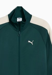 Dunkelgrüne Puma-Trainingsjacke mit Reißverschluss, hochgeschlossen, cremefarbenen Schulterakzenten und einem weißen Logo auf der linken Brust. Strukturiertes Material.