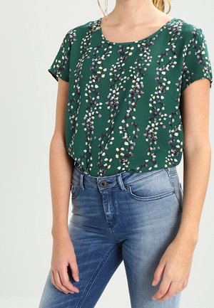 Kobieta nosząca zieloną krótką bluzkę w kwiatowy wzór, wpuszczoną w jasnoniebieskie jeansy z wysokim stanem, z przodem zapinanym na guziki i zamek błyskawiczny.