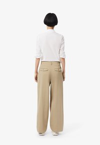 Camicia bianca con maniche arrotolate, abbinata a pantaloni beige a gamba larga. I pantaloni presentano due tasche posteriori con bottone.