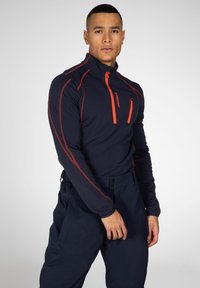 Navy blauwe fleece sweater met rits en oranje accenten en logo's. Heeft een hoge kraag en een getailleerd ontwerp, gecombineerd met donkere broeken. Glad textuur.