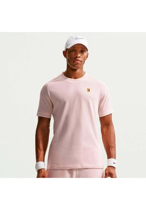 Férfi világos rózsaszín texturált rövid ujjú ingben, fehér Nike sapkában és fehér csuklópántokkal, egyszerű fehér háttér előtt áll.