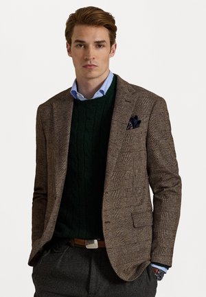 Brauner Hahnentrittmantel mit Spitzenrevers, getragen über einem grünen Zopfmustersweater. Hellbluse mit Kragen, graue Hose und ein blauer Einstecktuch.