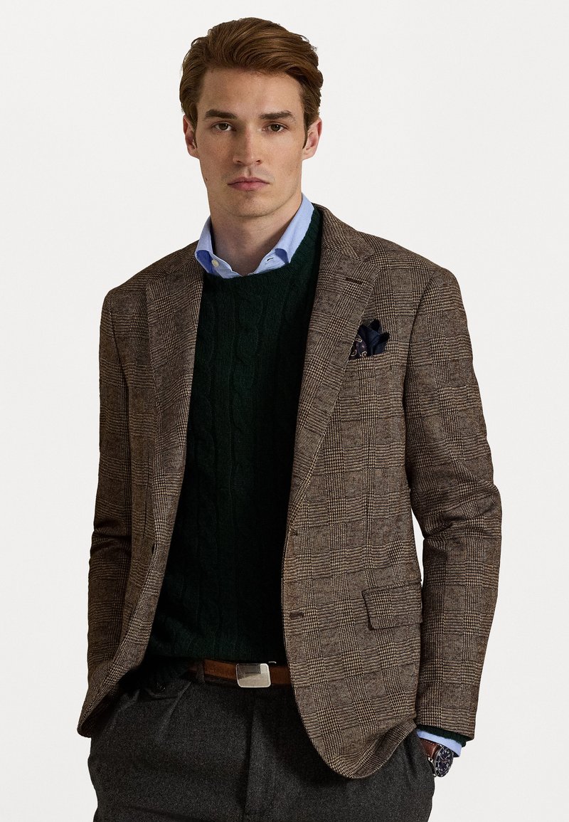 Blazer marrón de espiga con solapas en pico, combinado con un suéter de punto de cable verde. Camisa de cuello celeste, pantalones grises y un pañuelo de bolsillo azul.