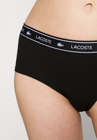 Fekete alsónadrág széles, elasztikus derékpánttal, amelyen a "Lacoste" márkaneve és logója látható. Simább pamut anyag, klasszikus rövid fazon.
