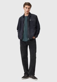 Marinblå jacka med en slät textur och en dragkedjeförsedd bröstficka, i kombination med en teal-färgad skjorta och svarta jeans. Modellen har på sig grå sneakers.