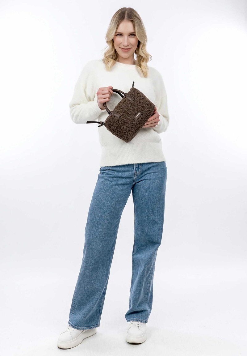 Bolso de mano de material de peluche marrón con asas cortas y acentos de cuero sintético, sostenido por una persona con un suéter blanco y jeans azules.