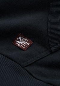 Czarny materiał dresowy o prążkowanej fakturze; posiada brązową tkana etykietę z białym napisem "DRY Super" i "EST 1954".