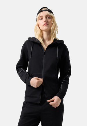 Jonge vrouw met blond haar, gekleed in een zwarte pet, een zwarte zip-up hoodie en bijpassende broek, die de ritssluiting met beide handen aanpast.