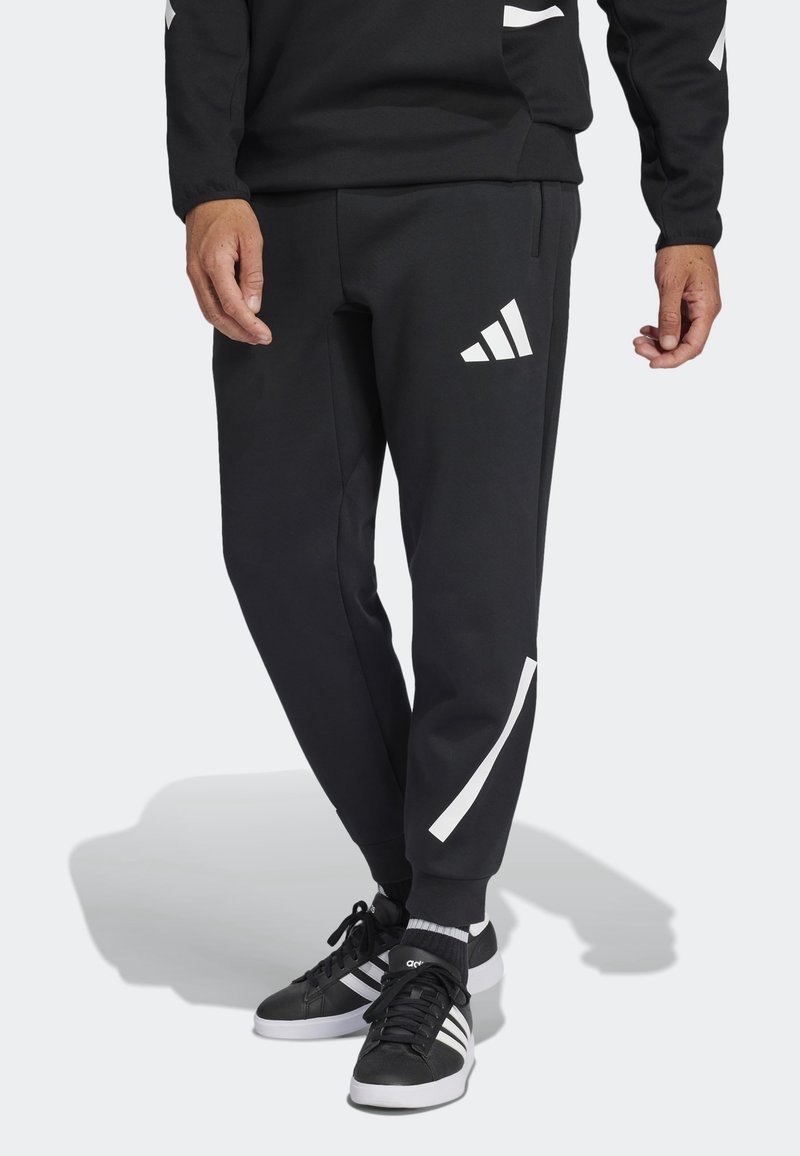 Underkropp av person som bär svarta Adidas sweatpants med vitt logo och ränder, ihop med svarta och vita Adidas sneakers.