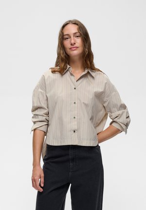 Vrouw draagt een beige, langärmelig, knoopshirt met dunne verticale strepen en zwarte hoge taille broek, staand met één hand op de heup.