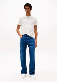 Hvit bomulls-t-skjorte, blå denim jeans med rett passform, hvite joggesko. Modellen står mot en enkel lys bakgrunn, med hendene i lomma.