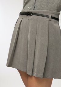 Primo piano di una gonna plissettata grigia con una sottile cintura nera indossata sopra una maglietta grigia con piccole decorazioni di perle.