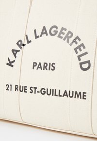 Sac en toile crème avec texte noir indiquant "KARL LAGERFELD PARIS 21 RUE ST-GUILLAUME", présentant des détails cousus et une forme rectangulaire.