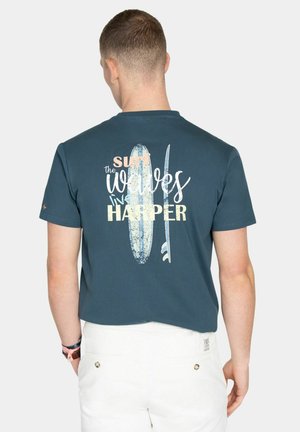 Hombre vestido con camiseta azul oscuro con gráfico de tabla de surf blanca y texto "Surf the waves live Harper" en la parte trasera, combinado con pantalones blancos.