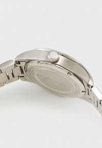 Nerezová ocelová hodinky s kulatým pouzdrem, hladkým rámečkem a texturovanou korunou. Obsahují kovový náramek s články a vyražené logo na zadní straně.