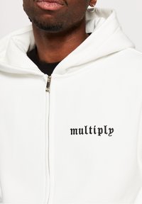 White zip-up hoodie with black "multiply" text on the chest. Features a hood, smooth texture, and metallic zipper. Simple design.
