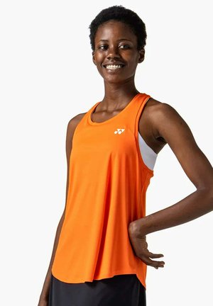 Mujer sonriente con cabello corto y rizado, vestida con una camiseta deportiva sin mangas naranja y pantalones negros, posando con una mano en la cadera.