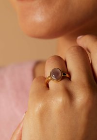 Gouden ring met een ronde, lichtpaarse steen, omrand door een parelaccent. Effen band, rustend op een zachte, getextureerde huidoppervlak.