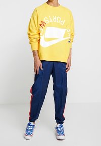 Felpa gialla con logo Nike bianco e lettering "SPORT" rosso. Pantaloni della tuta blu navy con strisce laterali rosse. Calzature con design blu e bianco.