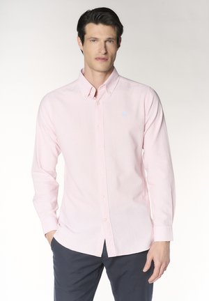 Uomo con camicia rosa chiaro abbottonata e pantaloni scuri, in piedi con una mano in tasca davanti a uno sfondo semplice.