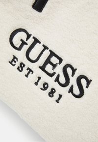 Tissu en molleton blanc cassé avec un texte brodé noir indiquant "GUESS EST 1981." Comprend un accent en cuir noir au-dessus du texte.
