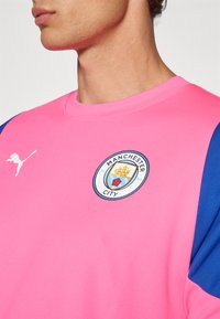 Αθλητική μπλούζα Puma με φωτεινό ροζ σώμα, μπλε μανίκια και έμβλημα της Manchester City. Λείο υλικό και στρογγυλή λαιμόκοψη.