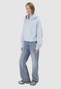 Młoda kobieta ubrana w jasnoniebieską bluzę z kapturem, luźne, wyblakłe niebieskie dżinsy i srebrne trampki, stojąca na neutralnym tle.