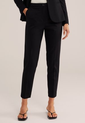Frau trägt schwarze, maßgeschneiderte Knöchelhosen, ein schwarzes Blazer und schwarze Riemchensandalen, steht auf neutralem Hintergrund.