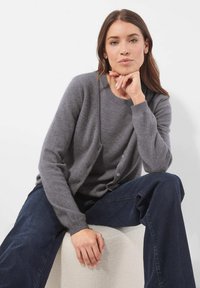 Frau mit braunen Haaren, die einen grauen Pullover und eine Strickjacke trägt, sitzt mit dem Kinn auf der Hand vor einem weißen Hintergrund.