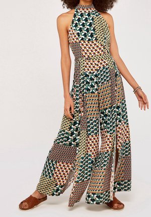 Kobieta ubrana w bezrękawnikową sukienkę maxi z dekoltem halter i kolorowe geometryczne wzory patchwork oraz brązowe plecione sandały na płaskim obcasie.