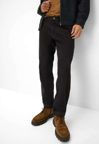 Zwarte katoenen broek met een rechte pasvorm, gecombineerd met bruine suède laarzen. De outfit omvat een donkere jas en een beige shirt eronder.